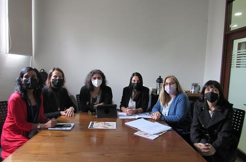 Un grupo de seis mujeres sentadas alrededor de una mesa en una reunión, todas usando mascarillas.