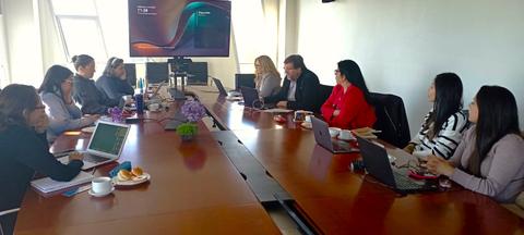 Una reunión de trabajo en una sala con varias personas sentadas en una mesa alargada y equipos tecnológicos presentes.