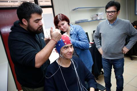 Un grupo de personas ajusta un electroencefalograma en la cabeza de un joven que sonríe mientras observa a su alrededor.