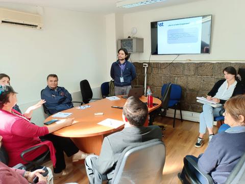 Una reunión en una sala de conferencias con varias personas sentadas alrededor de una mesa, mientras un presentador expone en una pantalla.