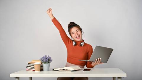 Una joven sonriente levanta el brazo con entusiasmo mientras trabaja en su computadora portátil en una mesa llena de libros y una planta.
