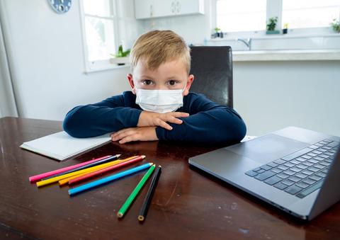 Un niño con mascarilla está sentado en una mesa frente a una computadora portátil y varios lápices de colores.