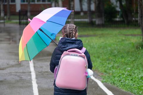Una niñita con trenzas lleva una mochila rosa y un paraguas colorido mientras camina por la calle.