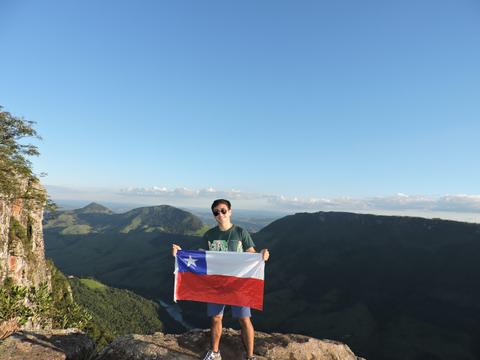 Un joven sostiene la bandera chilena en la cima de una montaña con un paisaje verde de fondo.