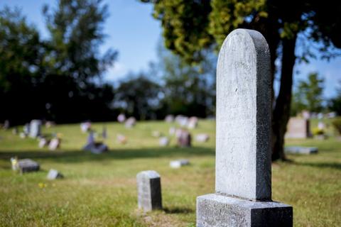 Una lápida de piedra se erige en un cementerio con varias tumbas al fondo, bajo un cielo despejado.
