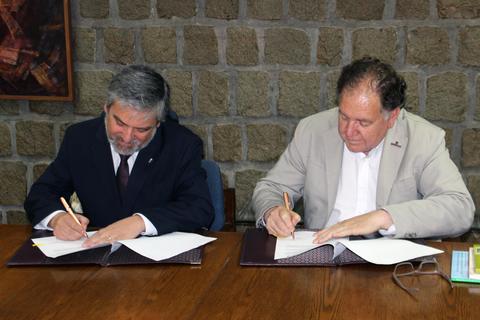Dos hombres firmando documentos en una mesa de madera.