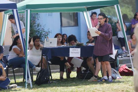 Un grupo de personas se reúne bajo carpas en un evento mientras una persona habla de pie frente a ellas.