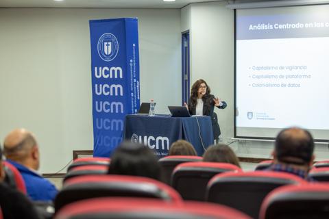 Una presentadora está exponiendo en un evento académico frente a una audiencia en un salón equipado con tecnología.