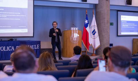 Un hombre está hablando frente a una audiencia en un auditorio, mientras se proyecta una presentación en una pantalla.