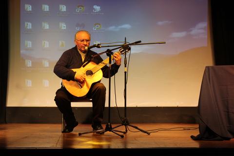Un hombre está sentado tocando la guitarra en un escenario con una pantalla de fondo.