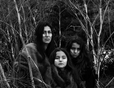 Tres mujeres posan en un entorno natural, rodeadas de ramas, con rostros pintados y atuendos oscuros.