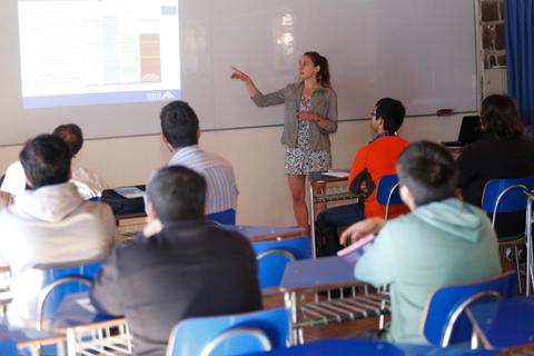 Una mujer presenta información en una pizarra frente a un grupo de estudiantes en un aula.