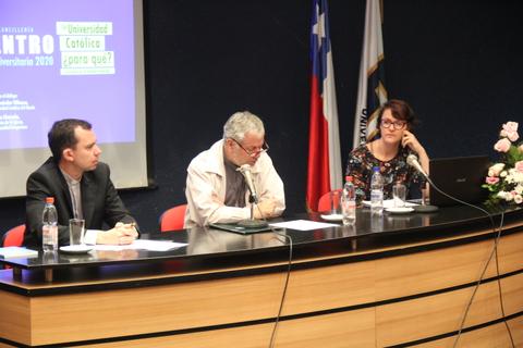 Una mesa de discusión en un evento universitario con tres panelistas hablando frente a una audiencia.