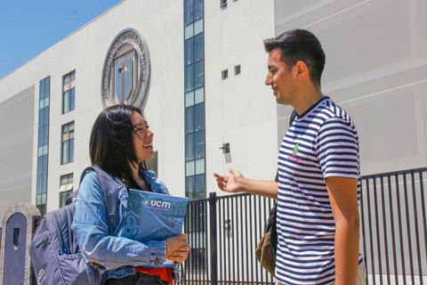 Dos estudiantes conversan frente a un edificio con el emblema de una universidad.