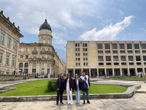 Un grupo de cuatro personas posa frente a dos edificios en una plaza con césped.