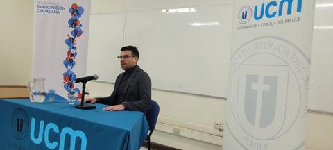 Un hombre sentado frente a un micrófono en una mesa con una tela de la Universidad Católica del Maule durante un evento.