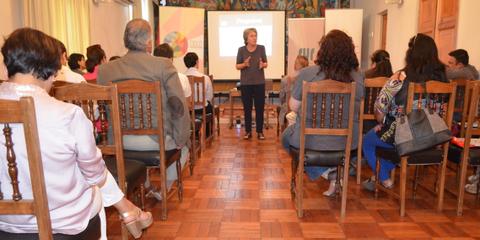 Una mujer está hablando frente a un grupo de personas sentadas en un salón.