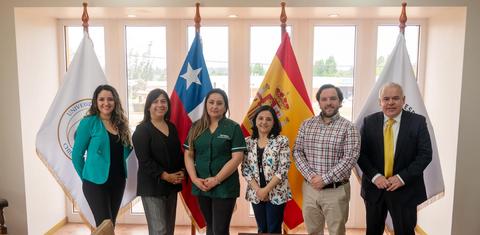 Un grupo de seis personas posando frente a banderas de Chile y España en un ambiente institucional.