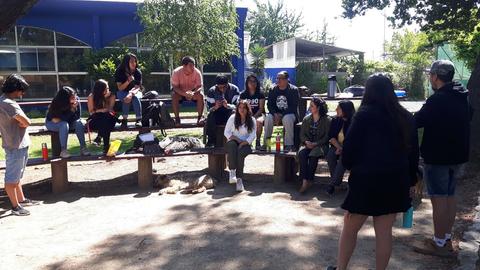 Un grupo de personas sentadas y de pie en un espacio al aire libre, participando en una actividad social.