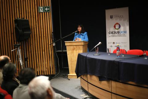 Una mujer habla desde un podio en un evento en un auditorio con asistentes en la sala.