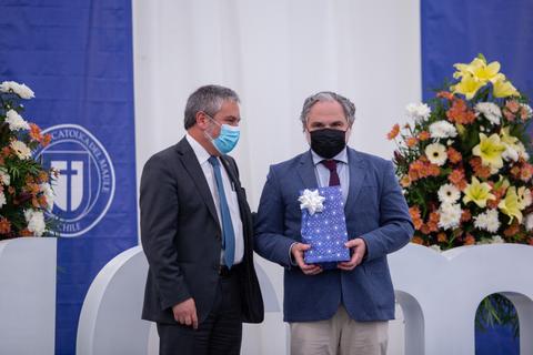 Dos hombres se encuentran en un evento, uno de ellos sostiene un regalo mientras el otro lo observa.