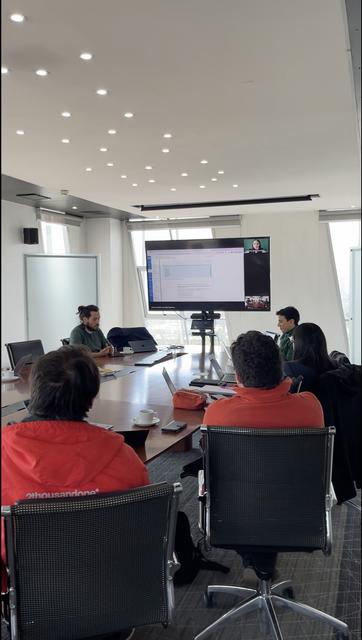 Una reunión en una sala de conferencia con personas sentadas alrededor de una mesa y una pantalla que muestra a alguien en videollamada.