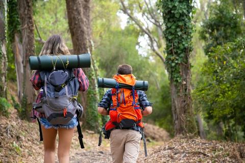 Dos personas caminan por un sendero forestal llevando mochilas y esterillas de camping.