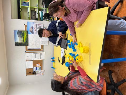 Un grupo de tres personas interactúa alrededor de una mesa amarilla mientras juegan con objetos de plástico de colores.