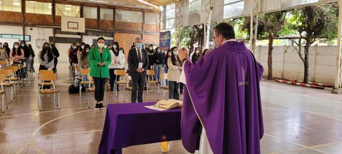 Una ceremonia religiosa con un sacerdote al frente y un grupo de personas en un auditorio escolar.