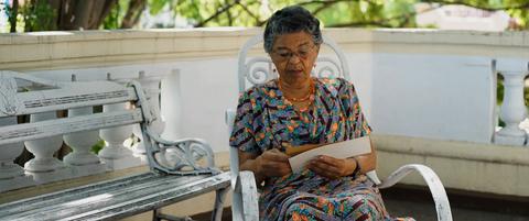 Una mujer mayor sentada en un banco, leyendo una carta con atención.