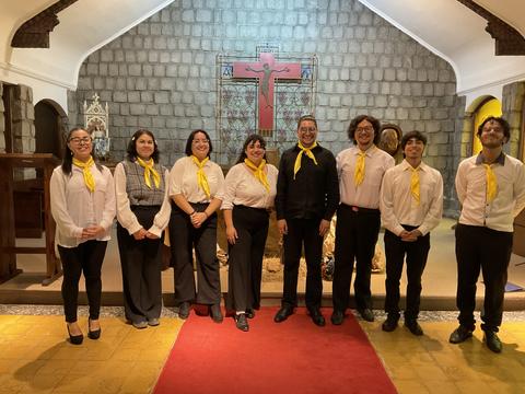 Un grupo de ocho personas vestidas de blanco y negro con pañuelos amarillos posan en un lugar con decoración religiosa.