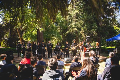 Un grupo de músicos se presenta en un evento al aire libre rodeado de árboles y espectadores.