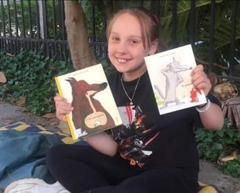 Una niña sonriente sostiene dos libros ilustrados en un entorno al aire libre.