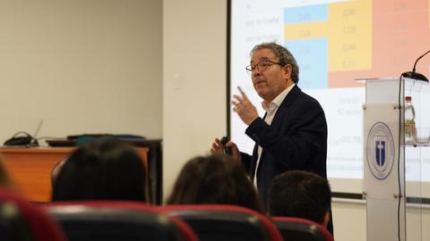 Un profesor está dando una charla en una sala de clases mientras muestra una presentación en pantalla.