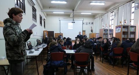 Un grupo de estudiantes en un aula escucha a un compañero que está leyendo un texto mientras un maestro observa en el fondo.