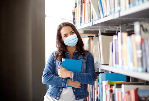 Una joven con mascarilla sostiene un libro en una biblioteca.
