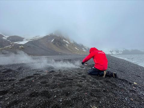 Una persona con chaqueta roja está trabajando en una playa de arena oscura y neblinosa junto al mar.