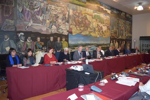 Una reunión formal con varias personas sentadas alrededor de una mesa, frente a un mural artístico.