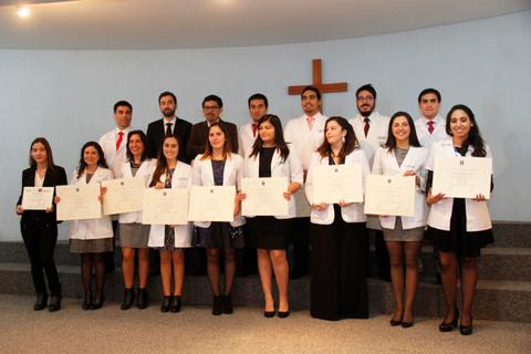 Un grupo de graduados sostiene sus diplomas en una ceremonia de finalización con una cruz al fondo.