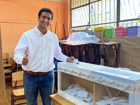 Un hombre sonriente muestra un pulgar hacia arriba mientras deposita un voto en una urna en un aula.