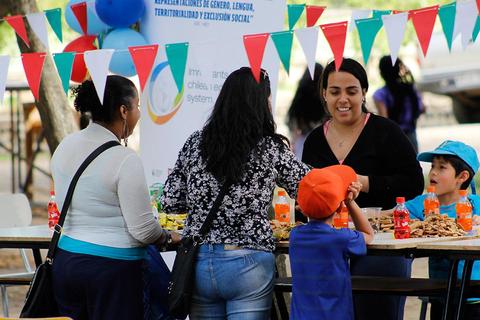 Un grupo de personas interactúa en una mesa llena de comida y bebida en un evento comunitario.
