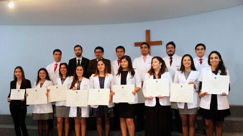 Grupo de graduados en acto de entrega de diplomas con un fondo de cruz en la pared.