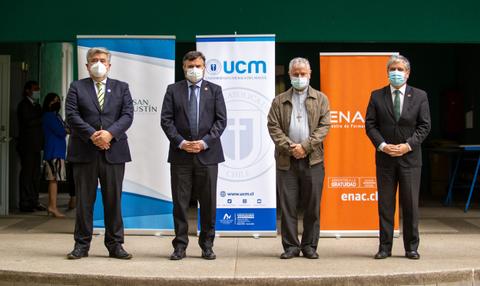 Cuatro hombres de pie frente a banners de instituciones educativas, todos usando mascarillas en un evento formal.