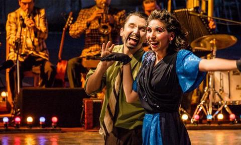 Una pareja de bailarines sonrientes se presenta en un escenario con una banda de música de fondo.