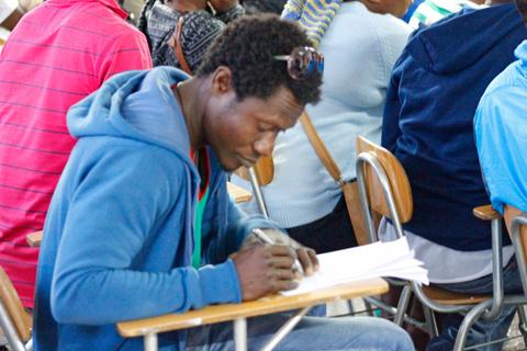 Un estudiante está escribiendo en una hoja en un aula con otros compañeros a su alrededor.
