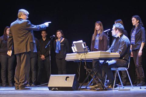 Un director de coro dirige a un grupo de cantantes mientras un pianista acompaña la interpretación.