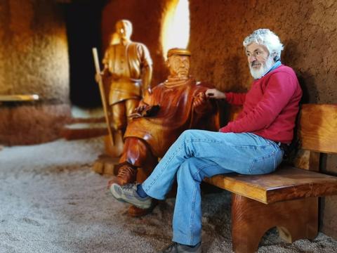 Un hombre con barba y suéter rojo se sienta en un banco junto a esculturas de madera de figuras históricas.