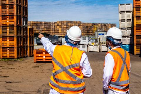 Dos trabajadores con cascos y chalecos reflectantes observan un área de almacenamiento llena de contenedores apilados.