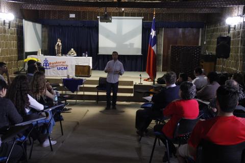 Un orador se dirige a un grupo de personas en una sala con banderas y un escenario.