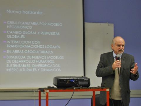Un hombre está presentando en una conferencia frente a una pantalla con texto sobre crisis planetaria y desarrollo humano.